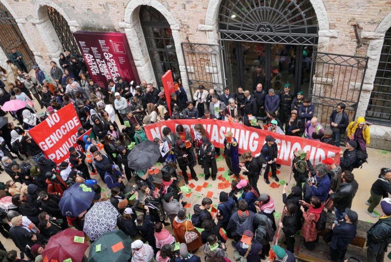 Devant l'entrée du pavillon du génocide lors de la Biennale de Venise, rassemblement et lecture de discours avec des banderoles : No Genocide Pavillon et No Artwashing Genocide. Des forces de l'ordre protège l'entrée et les représentantEs enferméEs dedans.